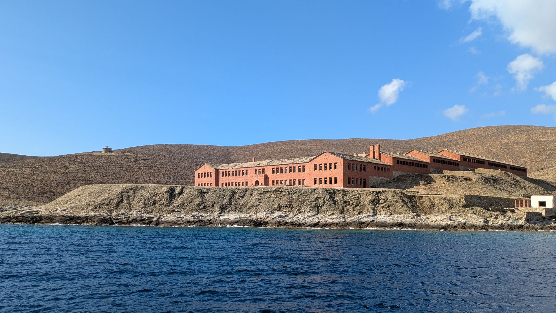 Picture of the main prison building in Gyaros, taken from onboard a boat. A clear sky with a white cloud in the corner is visible, as well as a deep blue calm sea